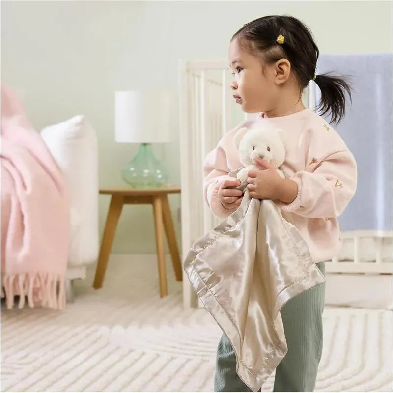 Child holding a Gund Spiritual Bear lovey, featuring a soft, white bear head and satin blanket, in a cozy room setting.