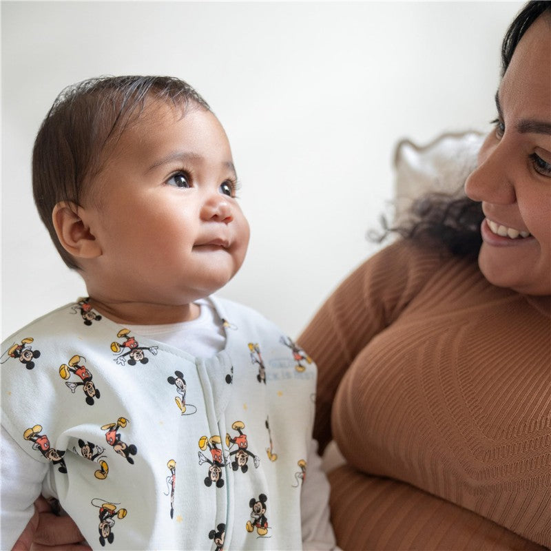 Infant wearing a HALO SleepSack Wearable Blanket featuring Mickey Mouse print, smiling at a woman holding them.