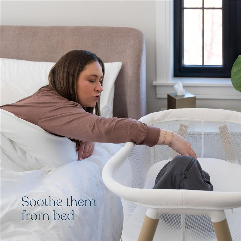 Woman reaching toward a HALO BassiNest Essential Sleeper, soothing her baby from bed, with soft bedding and natural light.