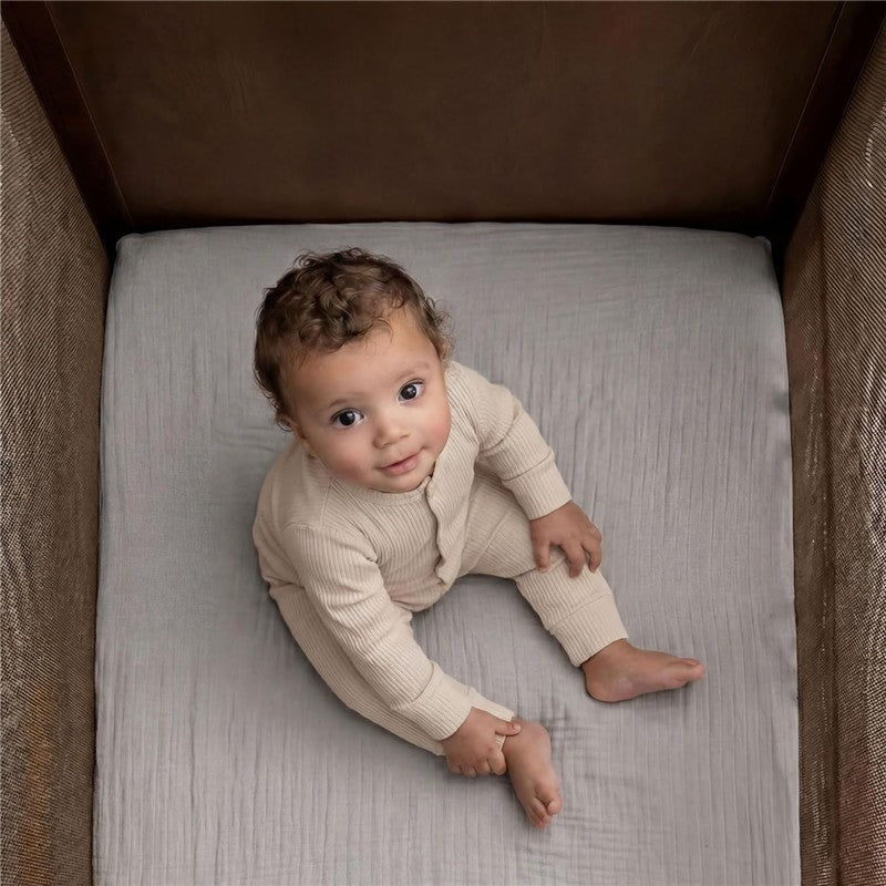 Full-size fitted crib sheets in storm grey with a baby sitting on one, looking up, dressed in a beige outfit.