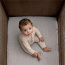 Full-size fitted crib sheets in storm grey with a baby sitting on one, looking up, dressed in a beige outfit.
