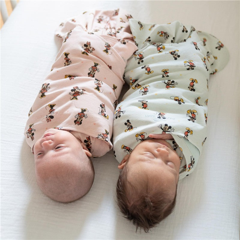 Two infants swaddled in HALO SleepSack Swaddle featuring Mickey Mouse designs, resting side by side on a light background.