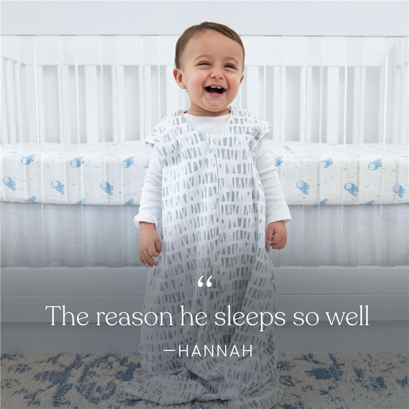 Baby wearing a HALO SleepSack Micro-Fleece blanket with a gray pattern, smiling next to a crib with whale-patterned bedding.