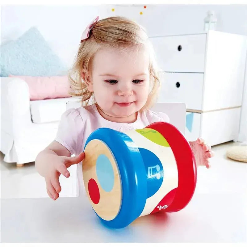 Toddler with a light pink shirt plays with a colorful Hape baby drum featuring blue, red, and green accents.
