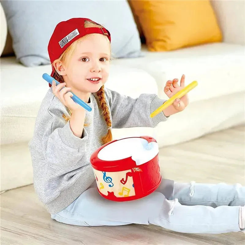 Child playing with a Hape electronic drum, holding blue and yellow drumsticks, smiling with visible musical notes on the drum.