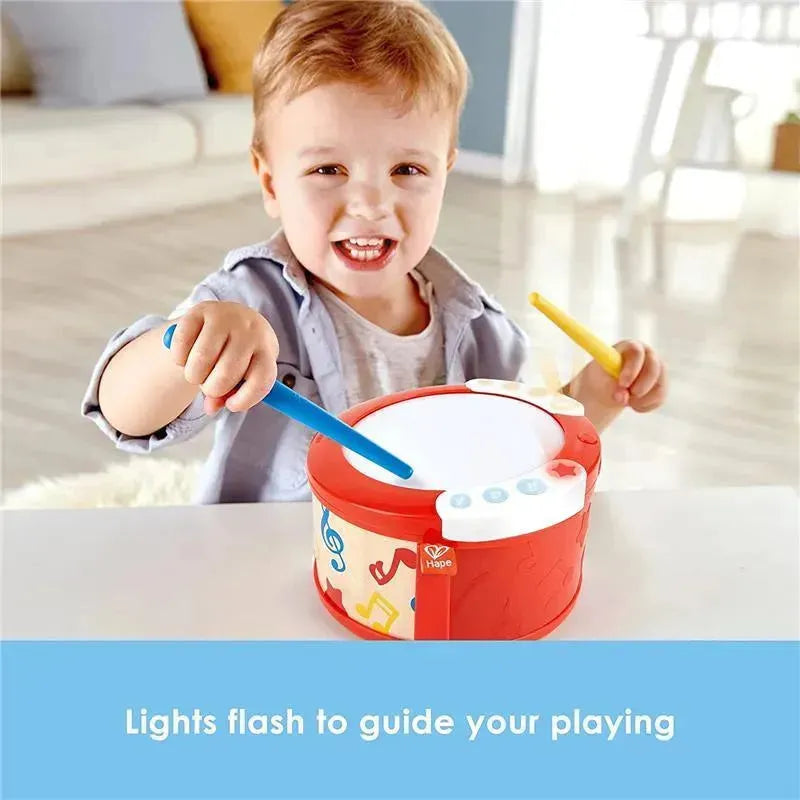 Young child playing with a Hape electronic drum, smiling while using blue and yellow sticks; drum features colorful musical notes and flashing lights.
