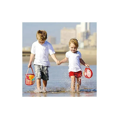 Two children, a boy and a girl, hold hands while playing in shallow water at the beach, holding a colorful bucket and a fish-shaped toy.
