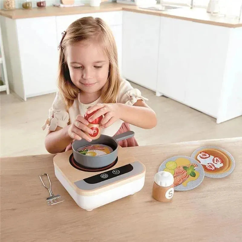 A child is playing with the Hape Fun Fan Fryer, pouring play food into a gray pot on a wooden tabletop stove.