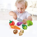 Child using a toy knife to slice colorful plastic vegetables, including a tomato, carrot, and broccoli, while smiling.