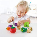 Child using a toy knife to cut colorful vegetable play pieces, including a red tomato, green broccoli, and mushrooms.