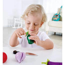 A young child with curly hair smiles while playing with Hape Garden Vegetables, holding a green toy vegetable and a toy knife on a table.