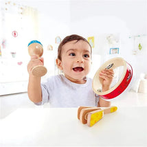A cheerful toddler holding a maraca and a tambourine, with wooden rhythm instruments on a table.