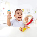 A cheerful toddler holding a maraca and a tambourine, with wooden rhythm instruments on a table.