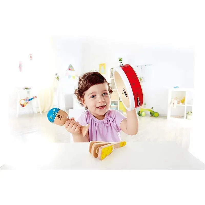 A young girl is joyfully playing with a Hape Junior Percussion Set, holding a red tambourine and a blue maraca, with wooden instruments on the table.