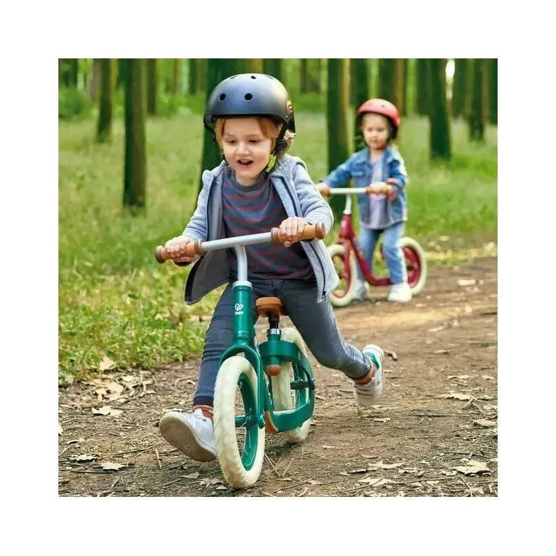 Two children riding balance bikes in a forested area; one bike is red, the other is teal.