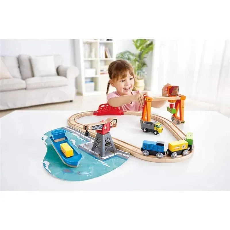 Child playing with the Hape Lift & Load Harbor Set, featuring a wooden train track, a harbor, and toy trucks and boats.