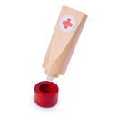 Wooden toy medical set piece resembling a tube of cream with a red cross symbol, tilted above a red circular base.