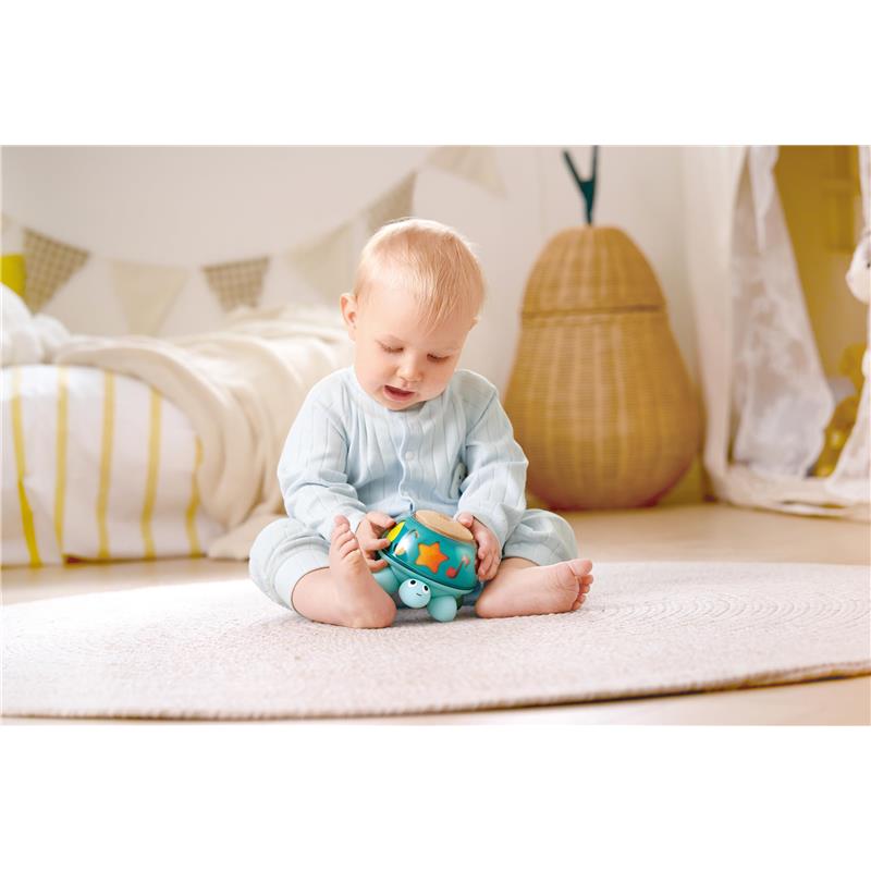 A baby in a light blue outfit plays with the Hape Music Turtle toy, featuring colorful musical elements, on a soft rug.