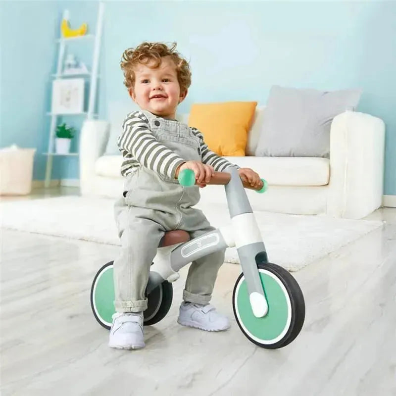 Child sitting on a green Hape balance bike indoors, smiling while holding the handlebars.