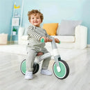 Child sitting on a green Hape balance bike indoors, smiling while holding the handlebars.