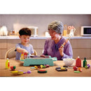 Young boy and grandmother play with the Hape Sizzling Griddle & Grill BBQ, showing colorful toy food items and sauces.