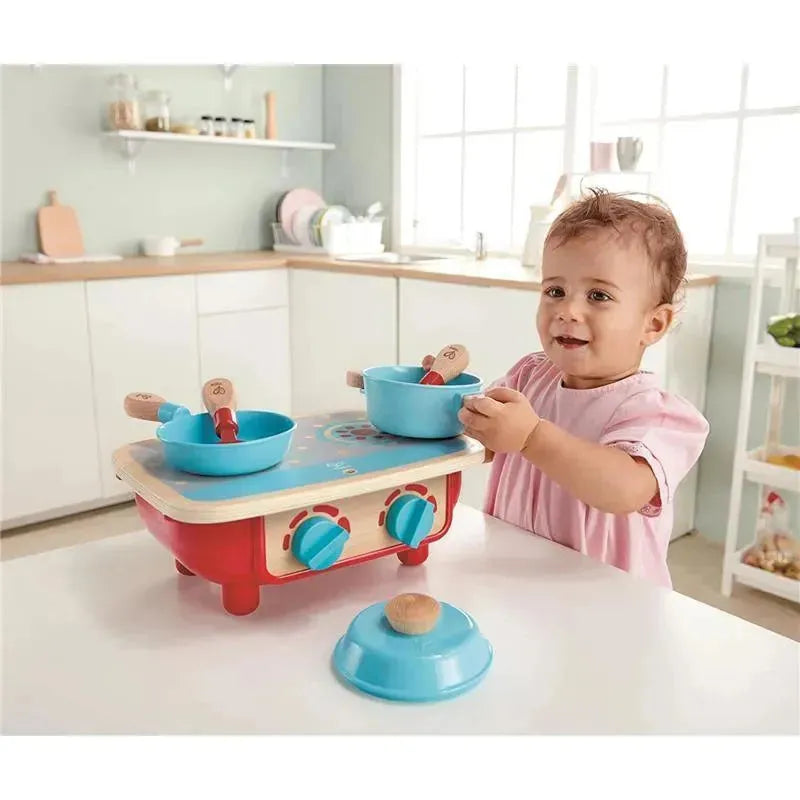 Toddler playing with a Hape kitchen set, featuring a red stove with blue bowls and wooden utensils in a bright kitchen.