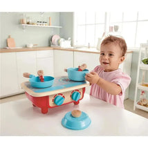 Toddler playing with a Hape kitchen set, featuring a red stove with blue bowls and wooden utensils in a bright kitchen.