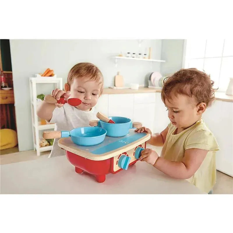 Toddlers playing with a Hape kitchen set, featuring blue bowls and wooden utensils on a red stovetop.