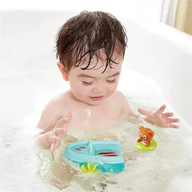 A child splashing in a bath plays with a blue pull-back boat toy and a small floating bear on a green ring.
