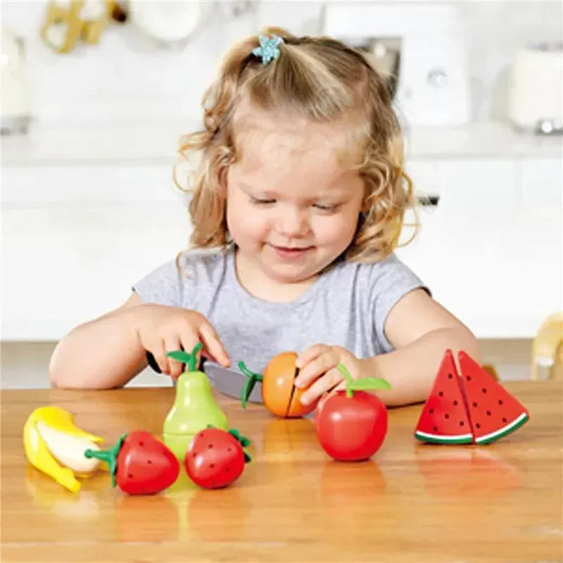 Child smiling while playing with wooden toy fruits, including bananas, strawberries, and a watermelon, using a play knife.