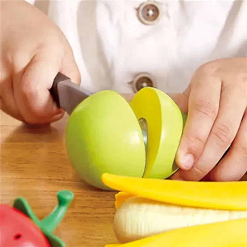 Child using a play knife to slice a green wooden apple, surrounded by other wooden fruits, including corn and yellow bananas.