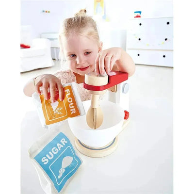 A girl is using a wooden toy mixer, adding flour and sugar packets into a bowl, engaging in imaginative play.