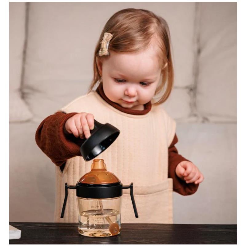 A toddler with light brown hair holds the lid of a black Heorshe dental-care sippy cup with orange accents, inspecting it.