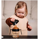 A toddler with light brown hair holds the lid of a black Heorshe dental-care sippy cup with orange accents, inspecting it.