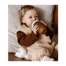Toddler sitting on a sofa, holding a Heorshe ultra wide neck baby bottle, wearing a beige outfit with a brown top.