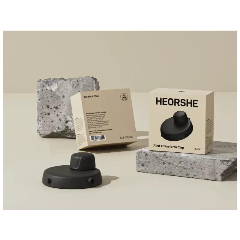 Black Heorshe Ultra Transform Cap displayed next to its packaging on a textured surface with concrete blocks.
