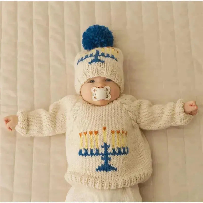 Light-colored knitted beanie hat and matching sweater with menorah design, worn by a baby sucking a pacifier.
