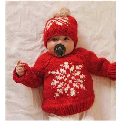 Baby wearing a red knitted crew neck sweater with a white snowflake design and a matching hat, lying down with a pacifier.