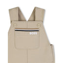 Infant cotton shortalls in stone color by Hugo Boss with adjustable straps and a front pocket featuring the logo.