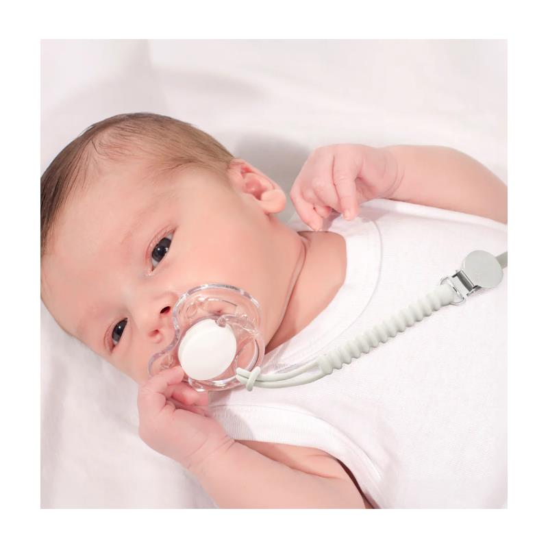 Baby with a clear pacifier attached to a black and white silicone holder, lying on a white surface.