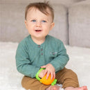Smiling baby sitting on a soft surface, holding an orange carrot-shaped teething mitt.