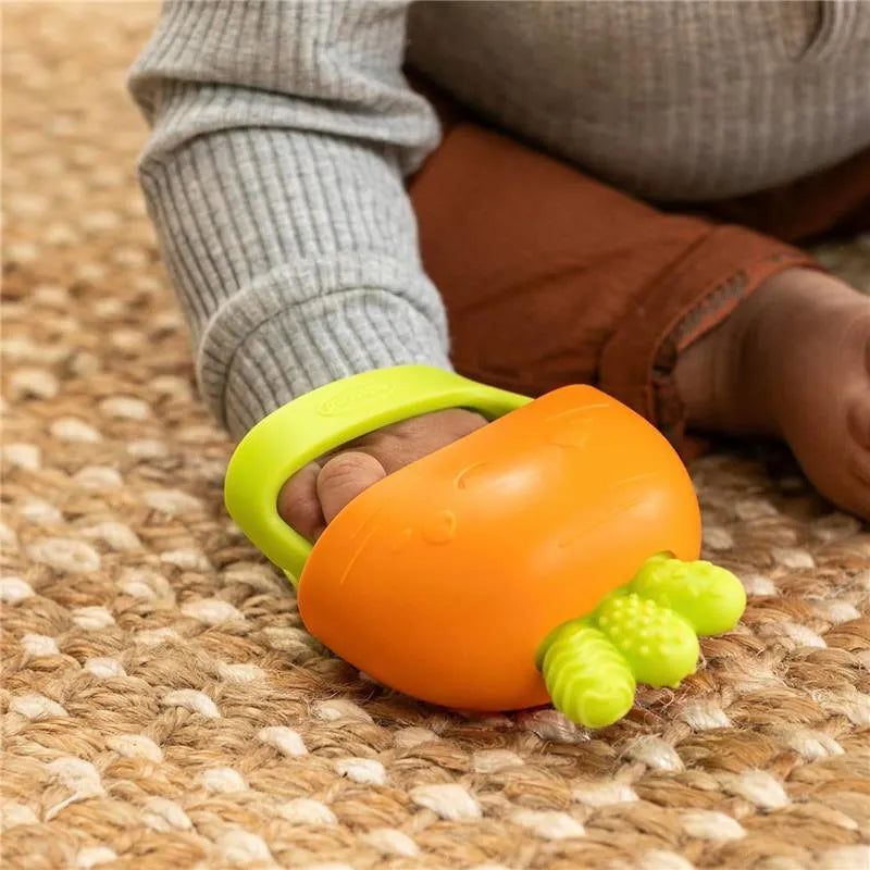Teething mitt designed like a carrot with a green handle held by a baby, resting on a textured surface.
