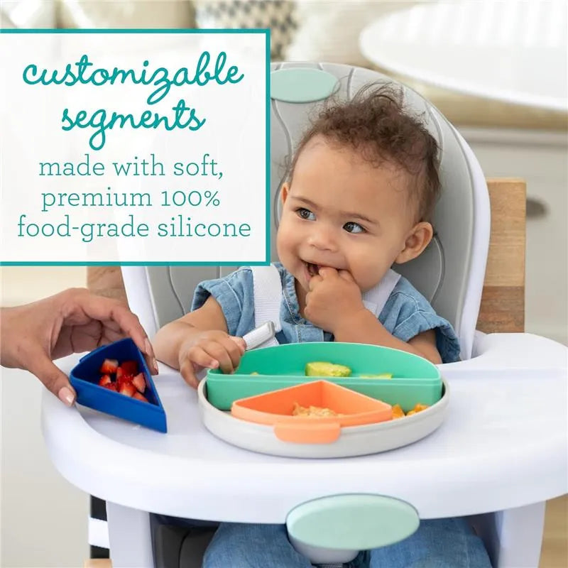 Highchair with a baby enjoying food from a colorful silicone plate featuring customizable segments and a hand offering fruit.