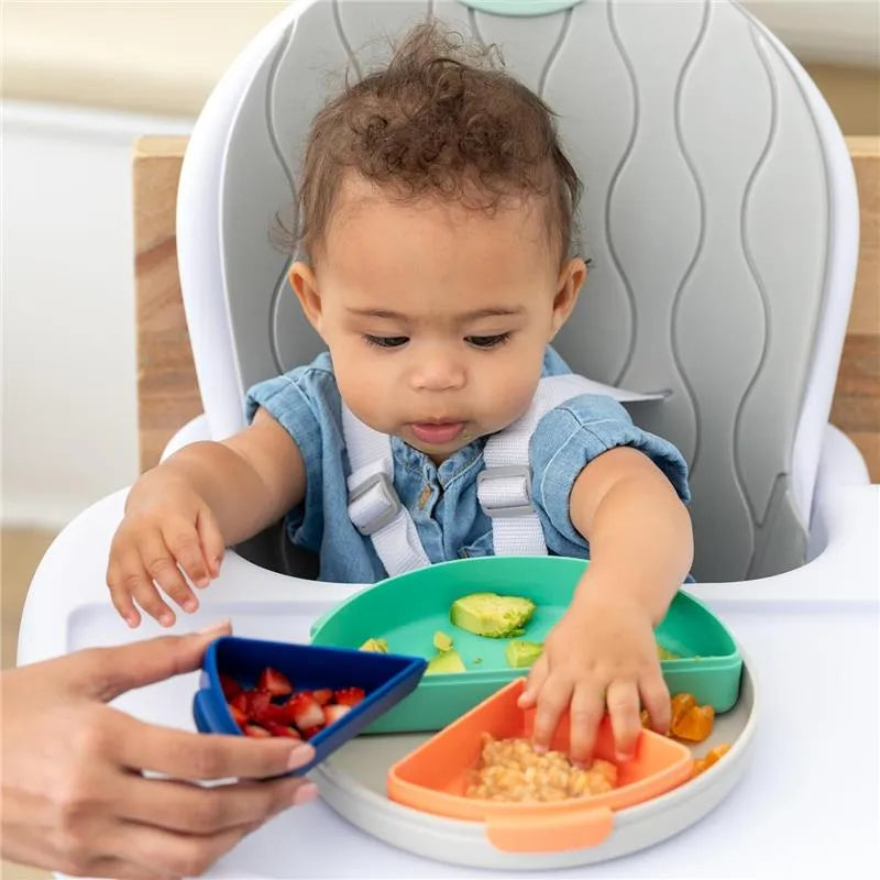 Toddler in a high chair engaging with a colorful segmented plate of food, including fruit and vegetables, assisted by an adult hand.