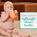 Creamsicle-colored silicone tumbler held by a toddler sitting on a carpet, featuring text about its lightweight and easy-to-hold design.