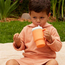 Child in a pink hoodie holds an orange silicone tumbler with a straw, sipping while sitting on a blanket outdoors.