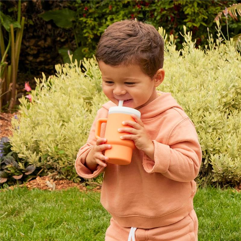 Toddler smiling while holding an orange Infantino My 1St Tumbler with a straw, standing in a garden.