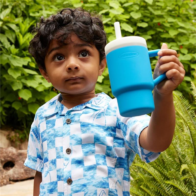 Child holding an Infantino My 1st Tumbler, a soft silicone 9 oz cup in ocean blue, surrounded by green foliage.