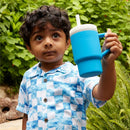 Child holding an Infantino My 1st Tumbler, a soft silicone 9 oz cup in ocean blue, surrounded by green foliage.