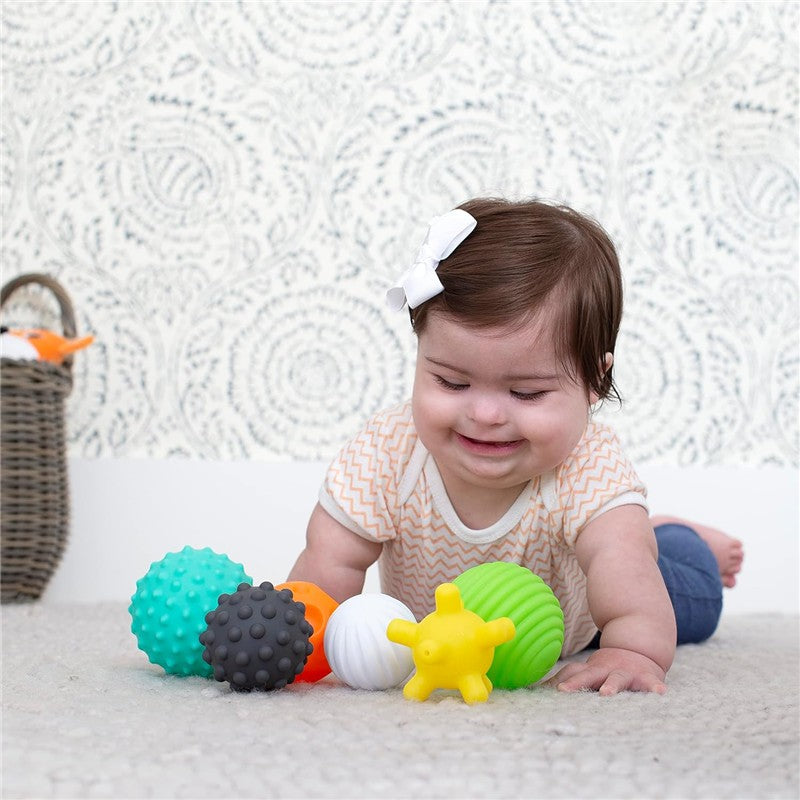 Multicolor Infantino Textured Multi Ball Set arranged around a smiling baby girl, who is playing on a soft surface.